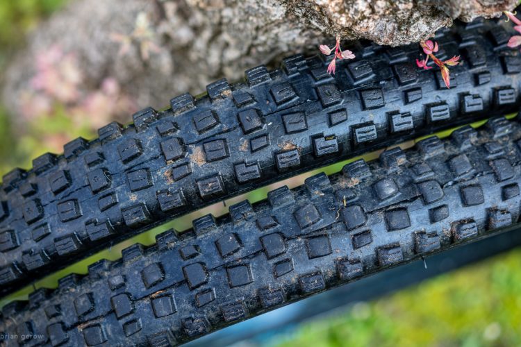 Close-up view of two bicycle tires stacked on top of each other, showcasing their textured tread patterns. The tires are slightly dirty, indicating use, and they are positioned near a tree with some small pink flowers visible in the background.