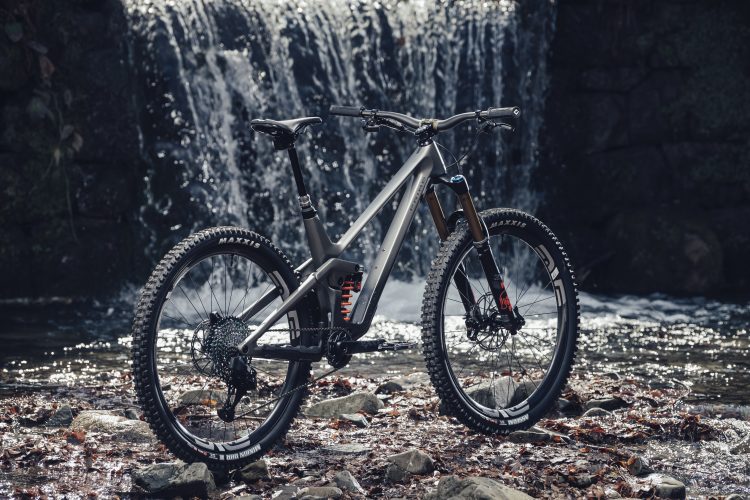 A modern mountain bike positioned on rocks by a serene waterfall, surrounded by autumn leaves. The bike features a sleek gray frame, knobby tires, and a dual suspension system, highlighting its rugged design suitable for off-road adventures.