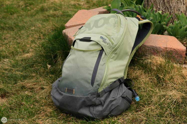A green hiking backpack resting on the grass, partially leaning against a stone. The backpack features a mesh front pocket and adjustable straps. The background includes patches of grass and a few stones.
