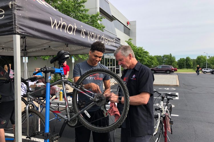 Two individuals are working together on a bicycle at an outdoor event under a tent. One person, with short curly hair, is focused on adjusting the bicycle's rear wheel, while an older man, wearing a black shirt, provides assistance. Tools and bicycle maintenance supplies are visible on a nearby table, and several bicycles are parked in the background.