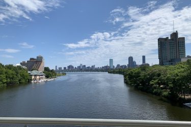 A scenic view of a river bordered by lush greenery, with a city skyline in the background under a clear blue sky. Several buildings, including a distinctive structure with a green roof by the water, are visible on the left, while taller skyscrapers line the horizon on the right. A few boats are seen on the river, contributing to the lively atmosphere of the area. Charles River Loop mountain bike trail.