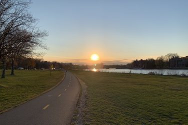 A scenic view of a sunset over a calm body of water, with a paved pathway winding through lush green grass. Silhouettes of trees and buildings line the shore, while the sun casts a warm glow across the sky, reflecting on the water's surface. Charles River Loop mountain bike trail.