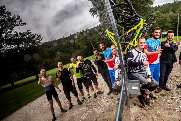 A group of mountain bikers posing for a photo in a natural setting. They are smiling and making gestures of camaraderie, with several bicycles stacked behind them. The background features greenery and a cloudy sky. The image reflects a sense of adventure and enjoyment in outdoor sports.