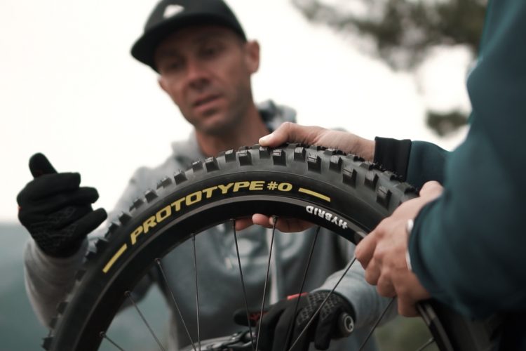 Two individuals are engaged in a discussion about a mountain bike tire, labeled "PROTOTYPE #0." One person, wearing a grey hoodie, is holding the tire and inspecting it, while the other person, wearing gloves, reaches out to examine the tire closely. The background features blurred natural scenery, suggesting an outdoor setting.