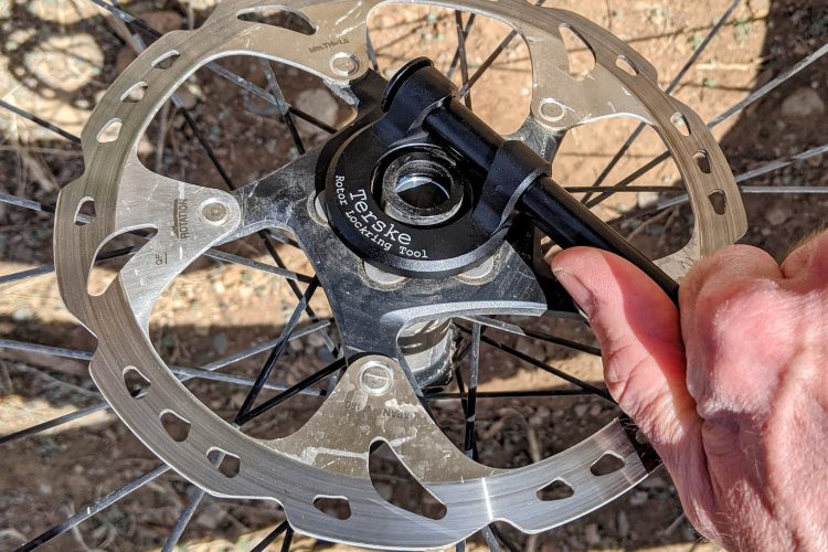 Close-up of a bicycle wheel's disc brake rotor, with a hand using a tool to adjust it. The background shows dirt and gravel, indicating an outdoor setting.
