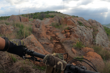 A view of a rocky mountain biking trail, featuring an assortment of large boulders and overgrown grass. The image captures the handlebar of a bike in the foreground, suggesting an active riding position. In the background, a green hill rises under a cloudy sky, with a glimpse of a body of water to the right. Lake Lawtonka Trails mountain bike trail.