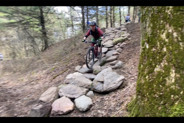 A mountain biker navigating a rocky trail through a wooded area, displaying skill and agility on a challenging section of the path. Rock Garden mountain bike trail.