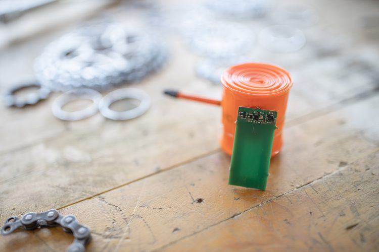 A close-up image of a green circuit board positioned in front of an orange coil of wire, with a blurred background featuring metal chain links and ring-shaped components on a wooden surface.