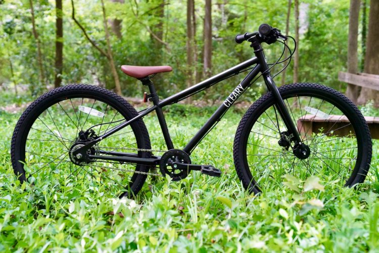A black bicycle with a brown seat resting on lush green grass in a wooded area. The bike features a simple design, suitable for outdoor riding, with a chain and textured tires. In the background, you can see trees and greenery, creating a natural, peaceful setting.