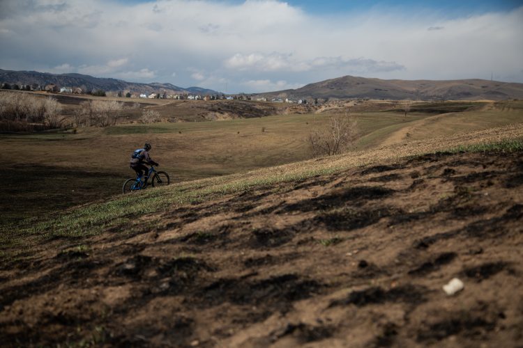 A cyclist riding a blue mountain bike along a grassy, sloped terrain under a partly cloudy sky. In the background, rolling hills and distant homes are visible, along with sparse trees and dry grass.