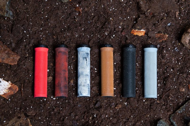 A row of six bicycle handlebar grips in various colors: red, brown, light gray, dark gray, black, and a weathered appearance. The grips are placed on a textured, dark soil surface, surrounded by small stones and wood fragments.