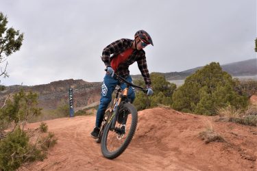 A person riding a mountain bike on a dirt trail, navigating a bend. The rider is wearing a helmet and a plaid shirt, with blue pants. In the background, there are hills and shrubs, under a cloudy sky. Red Fleet Downhill Flow Trail mountain bike trail.