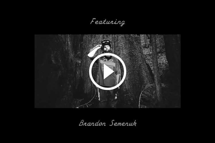 A black and white image featuring a mountain biker in front of a large tree, wearing a helmet and a jersey. The text "Featuring" is displayed at the top, while "Brandon Semenuk" appears at the bottom. A play button is centered within a circular border, indicating that it is a video.