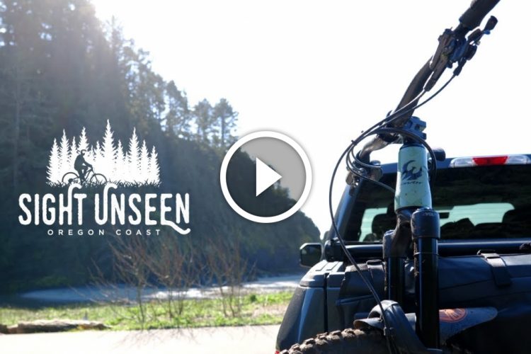 A scenic view of the Oregon Coast featuring a mountain bike mounted on the back of a truck, with lush trees in the background and the ocean visible in the distance. The image includes the logo "Sight Unseen" prominently displayed.