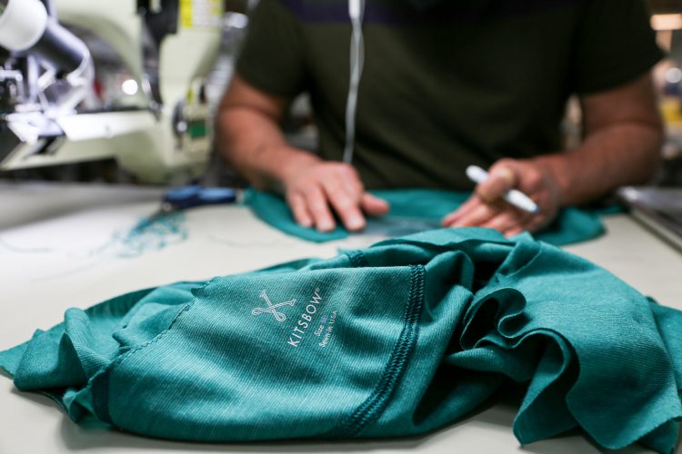A close-up of a green fabric with a logo that says "Kitsbow" lying on a work surface. In the background, a person is using a sewing machine and has their hands positioned on additional fabric, while a marker is placed nearby. The scene is set in a textile workshop.