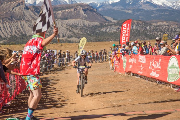 A mountain biker crosses the finish line at a race, while a flagger in a colorful shirt waves a checkered flag. Spectators line the course, cheering in the background with mountains visible in the distance under a clear blue sky. Red banners of sponsors are displayed along the race track.