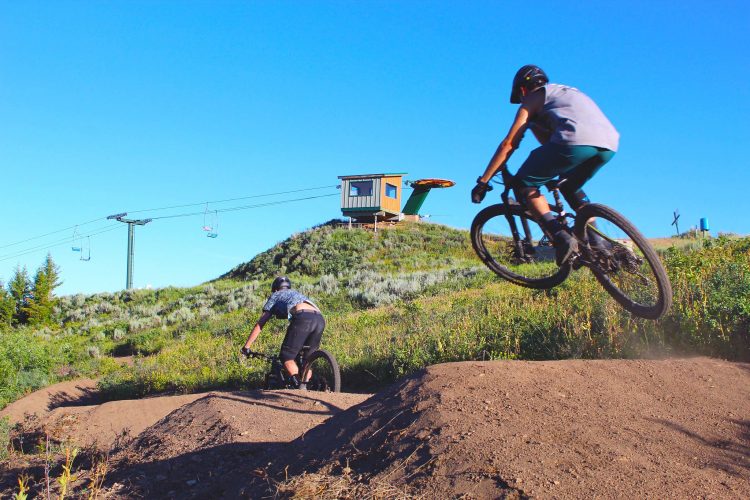 Two mountain bikers are riding on a dirt track against a backdrop of clear blue sky. One biker is airborne, performing a jump off a dirt mound, while the other is riding along the track. In the background, a ski lift and a small building are situated on a grassy hill. The scene is vibrant with green vegetation surrounding the biking area.