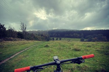 A mountain bike is positioned in the foreground, with its handlebars visible, looking out over a grassy field. The landscape includes rolling hills and a forest in the background under a cloudy sky. The scene suggests a remote, natural setting ideal for outdoor cycling. Vita slingan mountain bike trail.