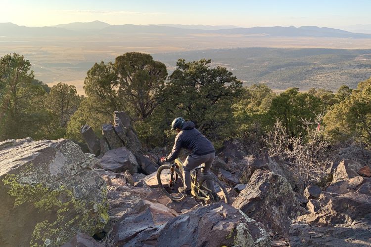 A person riding a mountain bike over rocky terrain on a hillside, surrounded by trees and with a panoramic view of a valley and distant mountains in the background. The scene is illuminated by soft morning light.