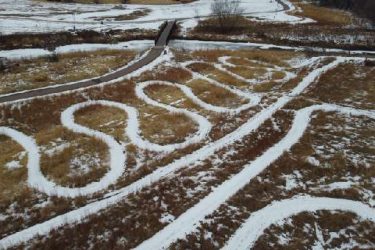 Aerial view of a snowy landscape featuring winding paths through golden grass, with a small wooden bridge in the background. The undulating trails create a rhythmic pattern across the terrain, contrasting against the patches of snow. Legacy Park mountain bike trail.
