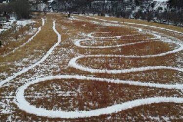Aerial view of a grassy field with winding snow-covered paths creating a meandering pattern through the tall grass. Legacy Park mountain bike trail.