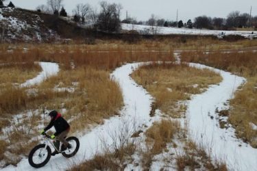 A cyclist riding a fat bike on a winding snowy trail through tall, brown grass in a winter landscape. The scene includes a hilly background and traces of snow on the ground. Legacy Park mountain bike trail.