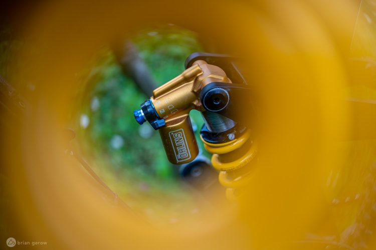 A close-up view of a gold coilover shock absorber, partially framed by a circular yellow object, set against a blurred green background. The shock absorber features adjustable components and branding details.