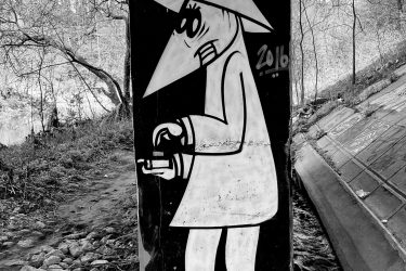 A black and white mural of a cartoonish figure with a large nose and a wide-brimmed hat, dressed in a coat and holding a small object in one hand. The mural is situated on a concrete structure in a natural setting, surrounded by trees and a rocky path. The year "2016" is visible on the artwork, along with some additional graffiti tags. Etobicoke Creek mountain bike trail.