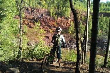 A mountain biker stands on a dirt trail surrounded by green trees, overlooking a rocky cliff with reddish hues in the background. The sun shines through the foliage, creating a serene outdoor scene. Redhead MTB Park mountain bike trail.