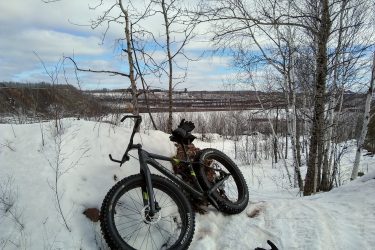 A black fat bike rests on a snowy trail, leaning against a pile of logs. In the background, a scenic view of a river and bare trees is visible under a partly cloudy sky. A black glove lies on the snow nearby, adding to the winter setting. Redhead MTB Park mountain bike trail.