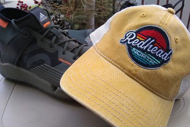 A close-up image of an Adidas trail shoe in black with orange accents placed next to a yellow cap featuring an embroidered design that reads "Redhead North." The background includes some greenery, suggesting an outdoor setting. Redhead MTB Park mountain bike trail.