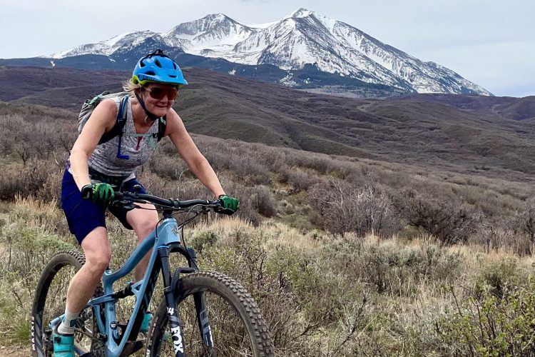 A person riding a mountain bike on a dirt trail, with snow-capped mountains in the background. The rider wears a blue helmet and sunglasses, and is dressed in a casual athletic outfit. Surrounding the trail are green grasses and shrubs, indicating a mountainous landscape in spring or early summer.