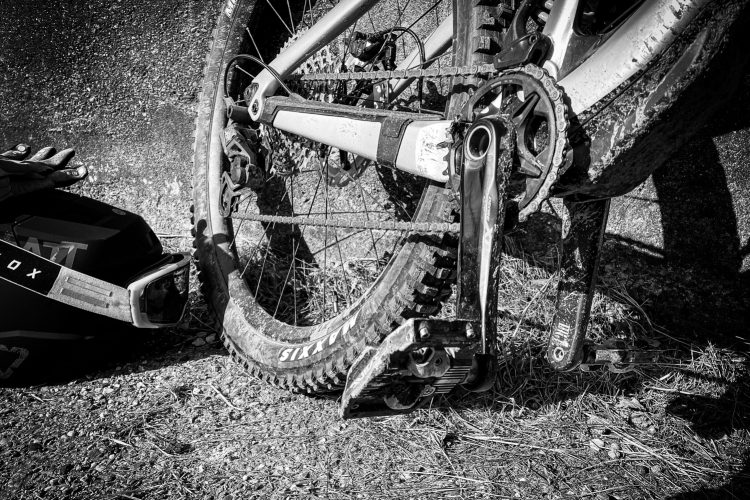 Close-up image of a mountain bike's rear wheel, crankset, and pedals resting on gravel. The bike shows signs of use with dirt and mud, and a pair of black gloves and a helmet are visible nearby. The image is presented in black and white.