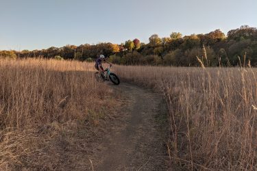 A person riding a mountain bike along a dirt path through tall, dry grass, surrounded by trees with autumn foliage. The scene is set in a natural landscape under a clear sky. Legacy Park mountain bike trail.