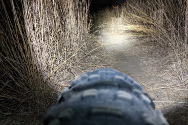 A close-up view of a tire on a narrow dirt path illuminated by a flashlight, surrounded by tall dry grass, under a night sky. Legacy Park mountain bike trail.