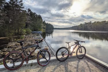 Two mountain bikes are parked on a gravel path near a serene lake, surrounded by trees. The sun is rising behind the clouds, casting reflections on the calm water surface, creating a peaceful outdoor scene. Wells Mills County Park mountain bike trail.