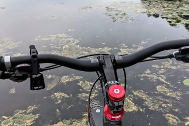 Mountain bike handlebars are positioned in the foreground, overlooking a tranquil lake with patches of lily pads and aquatic vegetation. The surrounding landscape features dense trees lining the water's edge under a cloudy sky. The bike is resting on a wooden pier or dock, creating a serene atmosphere ideal for outdoor activities. Wells Mills County Park mountain bike trail.