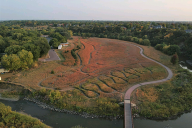 Aerial view of a riverbank landscape featuring winding pathways and a grassy area with reddish foliage. The scene includes trees in varying shades of green, a small wooden bridge crossing over the river, and suburban homes in the background, all under a soft, hazy sky. Legacy Park mountain bike trail.