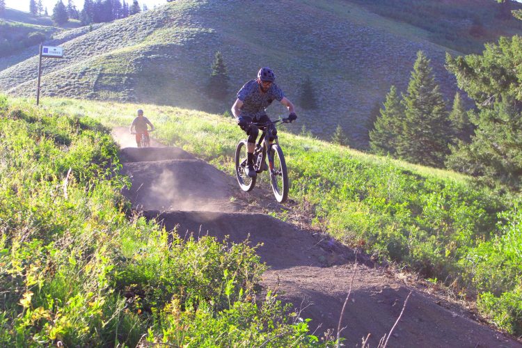 Two mountain bikers riding on a dirt trail in a lush, green landscape. One rider is airborne, jumping off a dirt ramp, while the other is visible in the background. The scene is set against a backdrop of rolling hills and trees under a clear sky. Dust is kicked up from the trail, indicating the dynamic movement of the bikers.