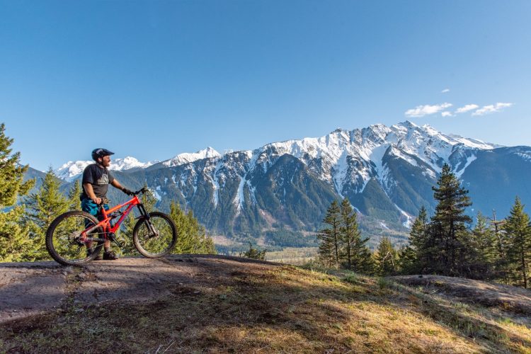 A mountain biker stands next to a bright red mountain bike on a rocky outcrop, overlooking a stunning mountain landscape with snow-capped peaks and a clear blue sky. Lush green trees surround the area, highlighting the natural beauty of the outdoors.