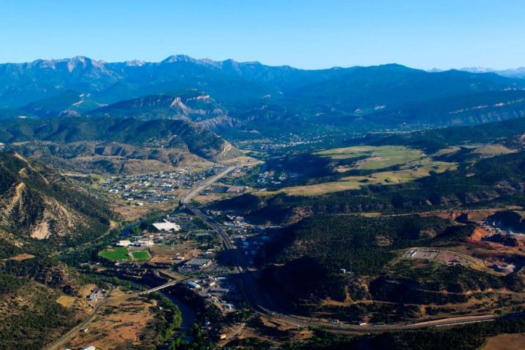 Aerial view of a mountainous landscape with lush green valleys, a winding river, and a small town nestled among the hills. The backdrop features majestic mountains under a clear blue sky.
