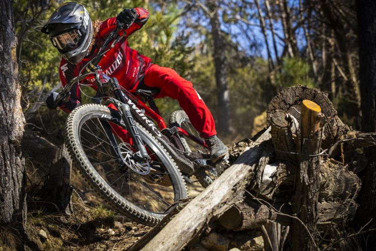 A person in red cycling gear maneuvers a mountain bike on a rocky trail, leaning into a turn near a log in a forested area with trees in the background. The rider wears a helmet and protective goggles, emphasizing the sport's adventurous nature.