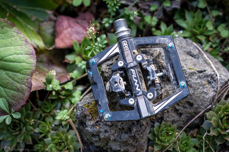 Close-up of a bike pedal resting on a rocky surface, surrounded by lush green foliage and colorful leaves. The pedal features a black frame with metallic accents and is partially covered in dirt, indicating outdoor use.