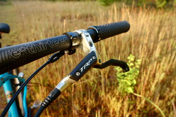 Close-up of a bicycle's handlebars, featuring a TRP brake lever and e*thirteen handlebar. The background consists of tall grass and a hint of greenery, suggesting an outdoor setting.