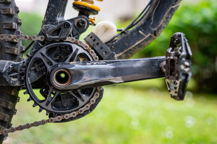 Close-up view of a mountain bike’s crankset, chain, and pedals, showcasing the intricate details of the drivetrain components against a blurred green background. The parts display signs of use, indicating the bike's adventurous history.