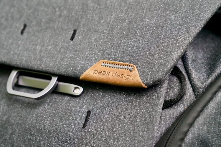 Close-up of a dark gray fabric bag featuring a leather tag embossed with "peak design" and a metal clasp for secure closure.