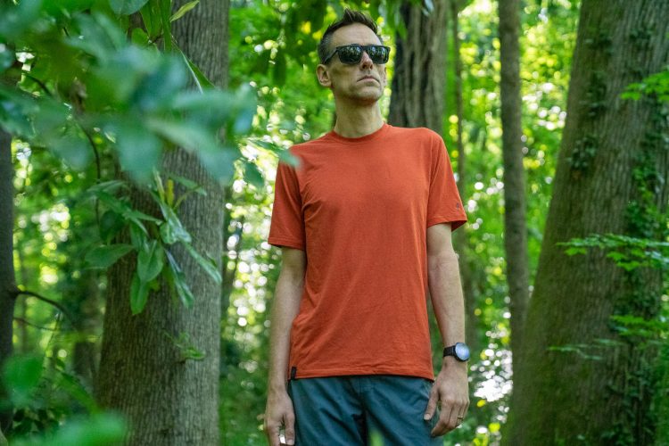 A man standing in a lush green forest, wearing sunglasses and an orange t-shirt paired with blue shorts. He is looking off into the distance, surrounded by trees and foliage. The sunlight filters through the leaves, creating a vibrant, natural backdrop.