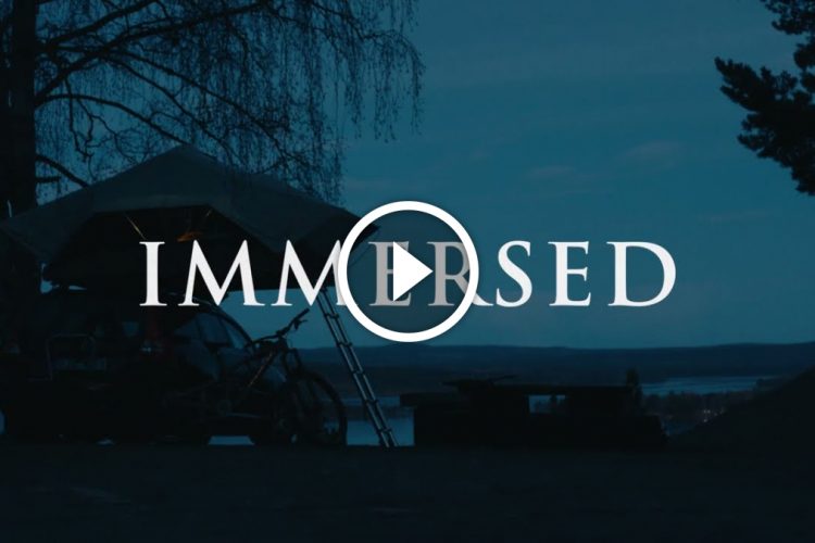 A serene nighttime scene featuring a tent atop a vehicle near a body of water, with the word "IMMERSSED" prominently displayed. A play button icon suggests a video is available, capturing the peaceful atmosphere of nature.