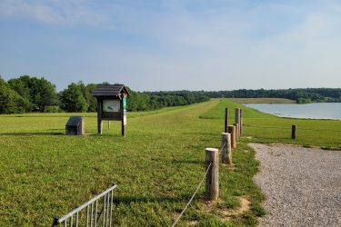 A scenic view of a grassy dam landscape with a walking path, featuring a wooden information kiosk and a stone marker in the foreground. The background includes a calm lake and lush trees under a clear sky. Wooden posts with a rope line the path leading towards the water's edge. McDougal Lake Trail mountain bike trail.
