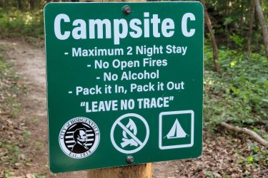 A green campsite sign reading "Campsite C," with guidelines including a maximum 2-night stay, prohibition of open fires and alcohol, and a reminder to "Pack it In, Pack it Out," along with the phrase "Leave No Trace." The sign features logos for the city of Hodgenville and symbols for no fires and camping. The background shows a wooded area. McDougal Lake Trail mountain bike trail.
